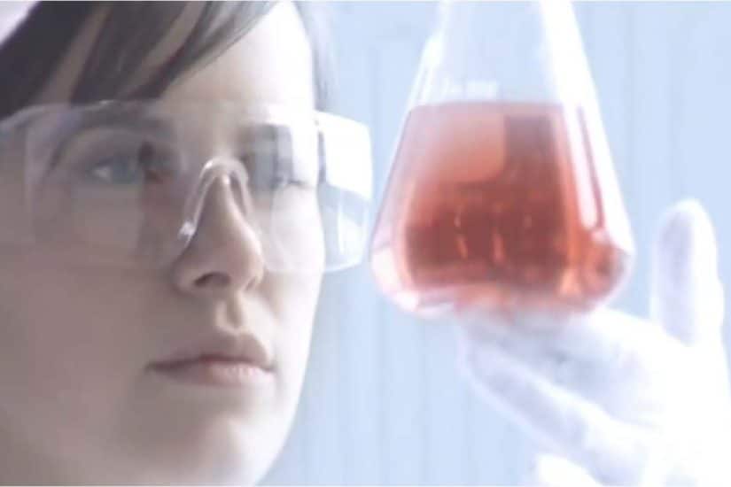 This is a photograph of a young woman examining a beaker full of red liquid.